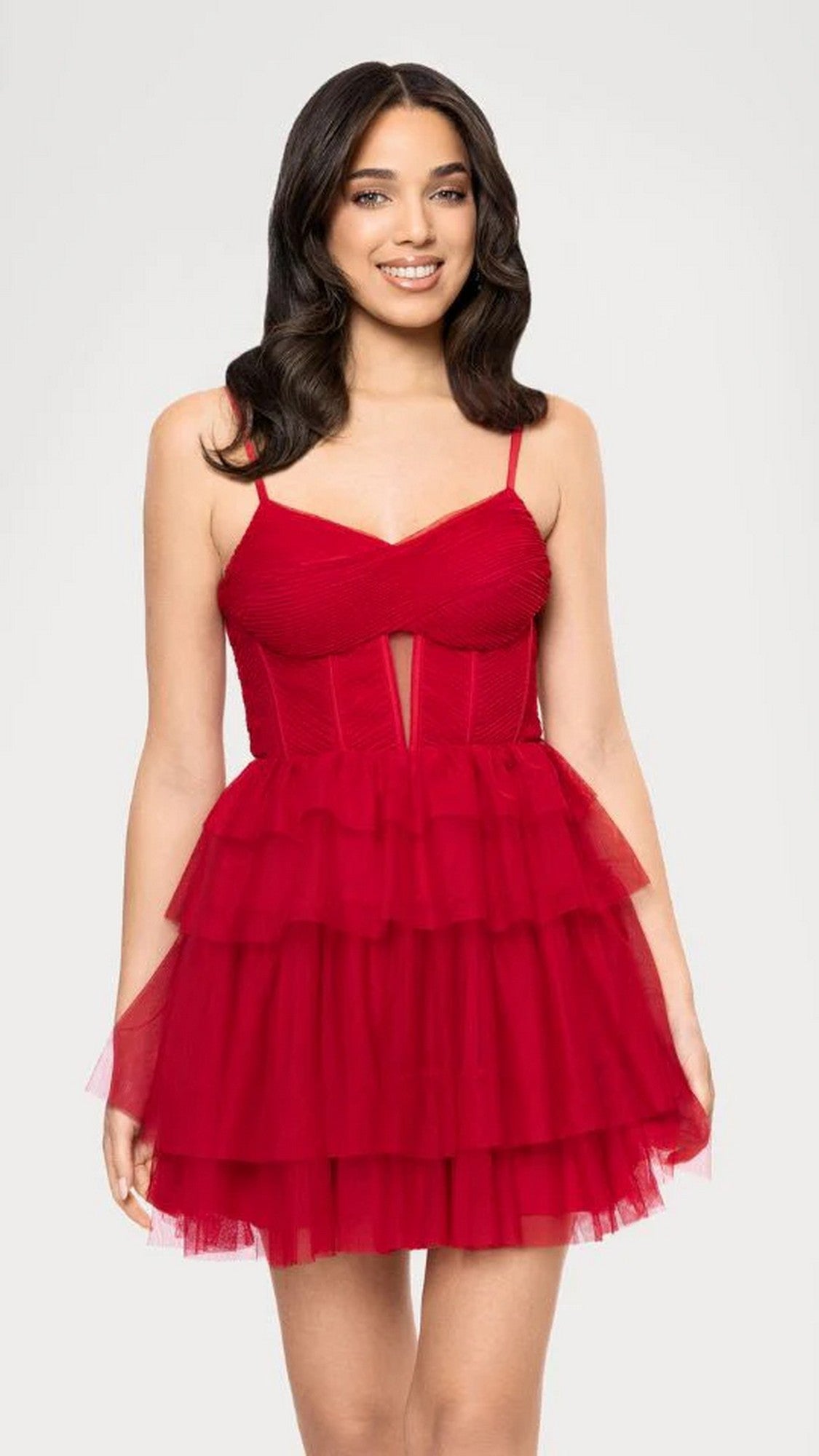 Woman stands smiling, facing forward, wearing the Blondie Nites 5842BN short red prom dress featuring a sleeveless fit-and-flare design with a ruched mesh corset bodice and layered tulle skirt against a light background.